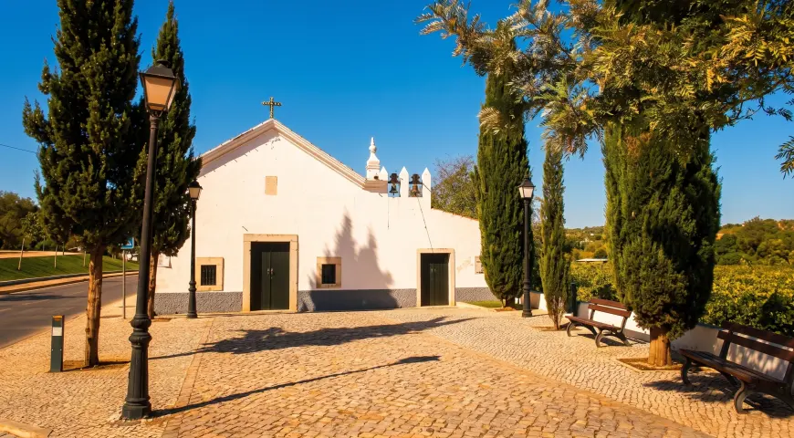 The low white Pé da Cruz chapel beside trees and a paved forecourt in Paderne