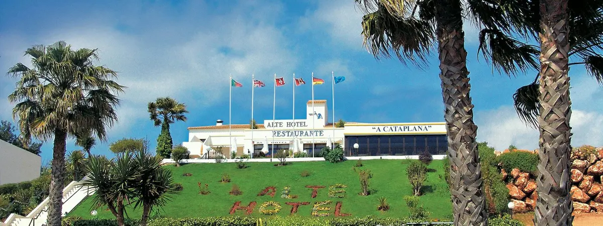 Alte Hotel’s white building, flags and landscaped approach beneath a blue sky