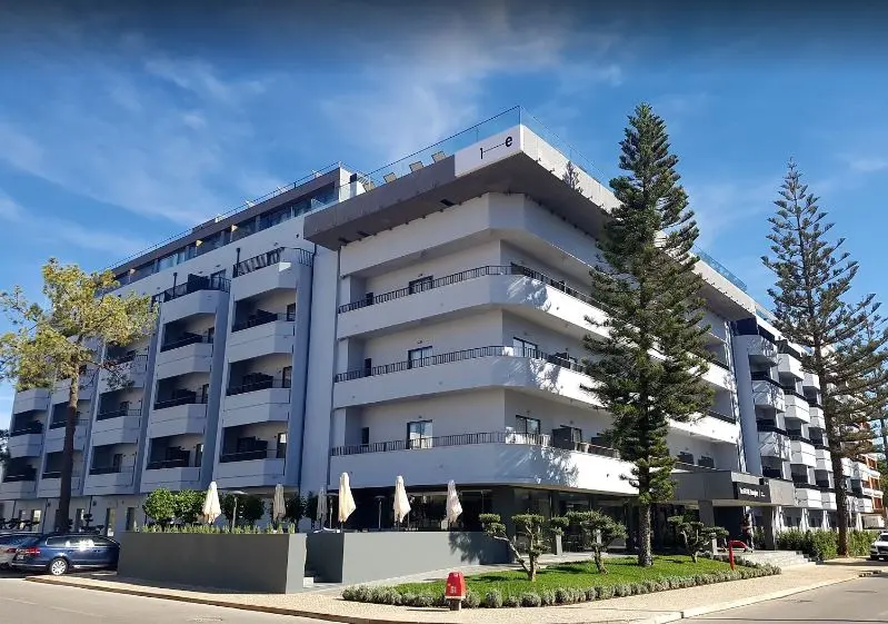 The pale balconies and street-corner entrance of The Prime Energize hotel between tall trees in Monte Gordo