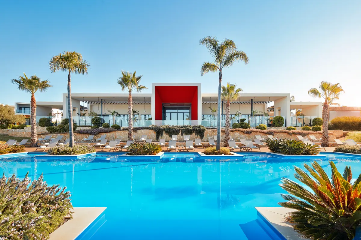 A broad blue swimming pool, palm trees and white low-rise resort building with a red entrance at Tivoli Alvor