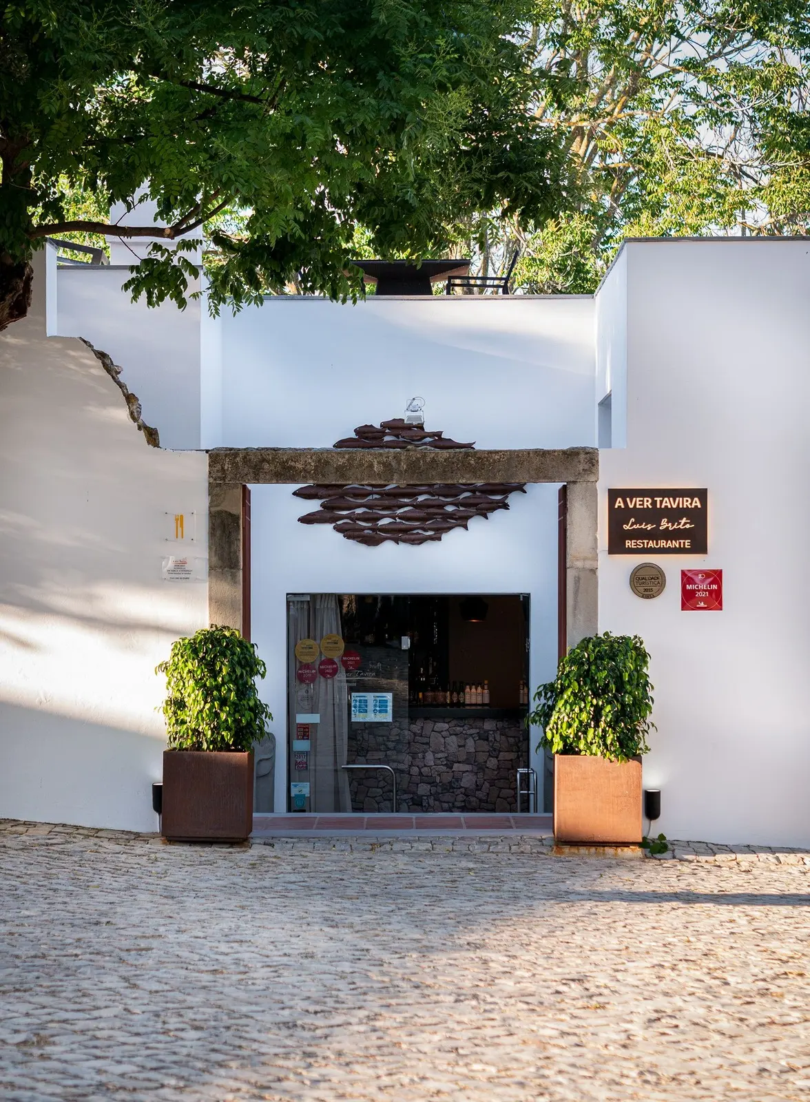 A Ver Tavira’s stone entrance and white walls beneath trees, with the restaurant name on the facade.