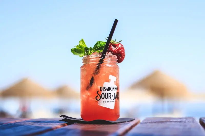 A red iced cocktail in a glass jar on a terrace table with beach umbrellas behind, from Borda d’Água’s gallery.