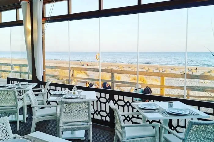 Tables beside the glazed sea-facing frontage of The Pr1me Beach Club in Monte Gordo.