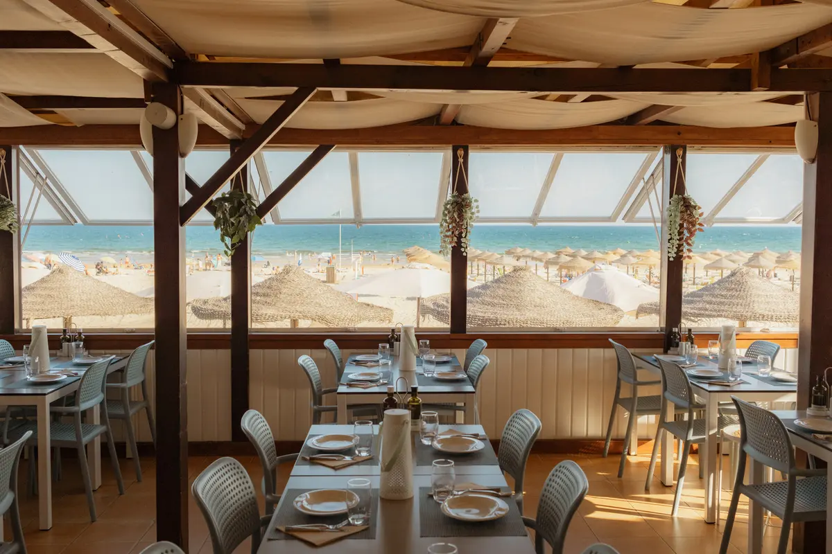 Set restaurant tables beneath timber beams looking towards Praia do Cabeço beach.