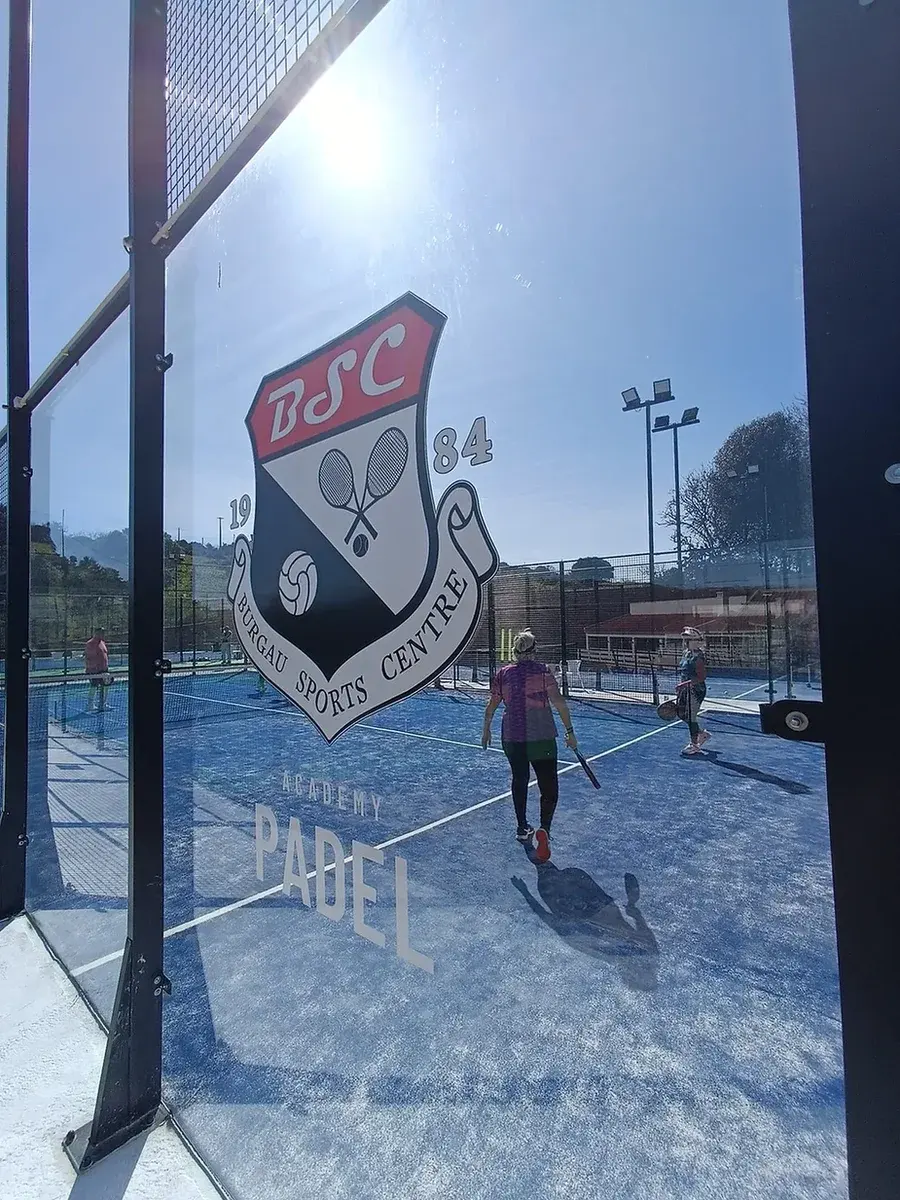 Players on a blue padel court behind Burgau Sports Centre branding printed on the court glass
