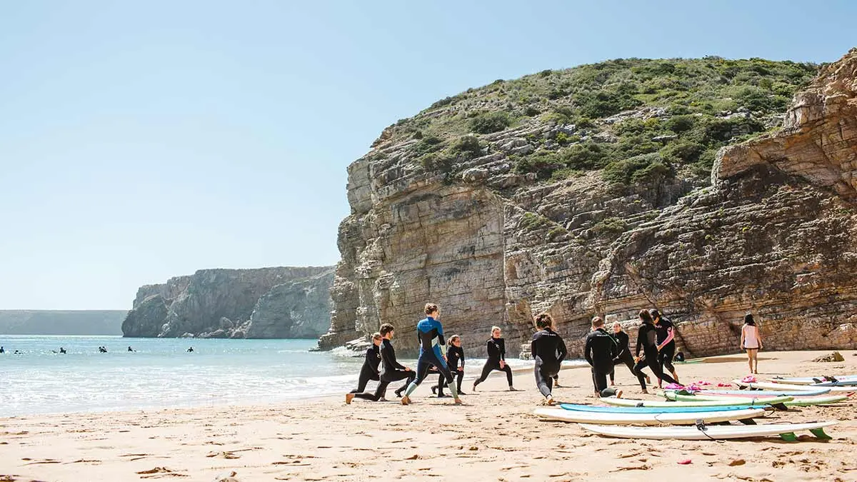 Students warming up beside surfboards on a beach beneath cliffs