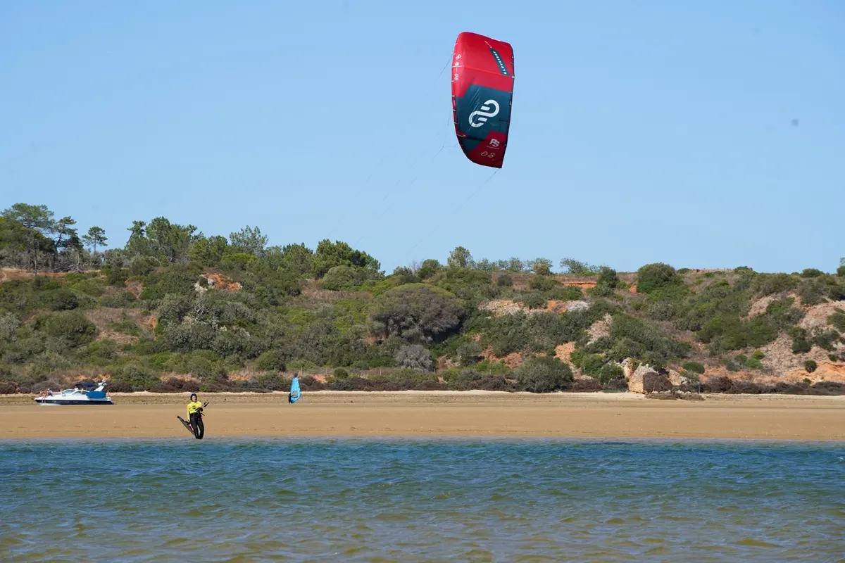 A rider handles a red kite beside the water in an Alvor Kite Center photograph
